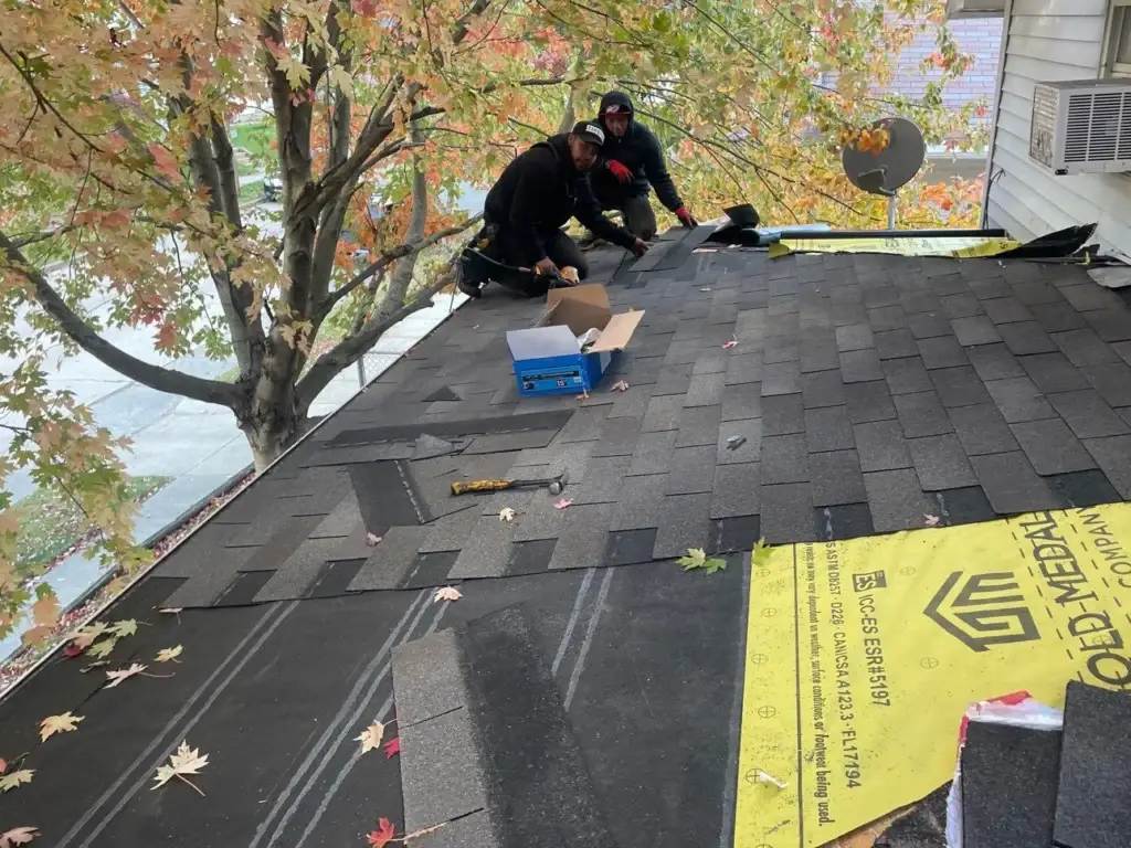 Two expert roofers perform repairs on a residential rooftop, surrounded by essential tools and materials. The roof features dark gray asphalt shingles, yellow underlayment and a satellite dish. A nearby tree boasts vibrant orange and yellow autumn leaves, contrasting with the industrial scene. The background reveals a suburban street with passing cars, showcasing the roofers' efficient work amidst daily life.