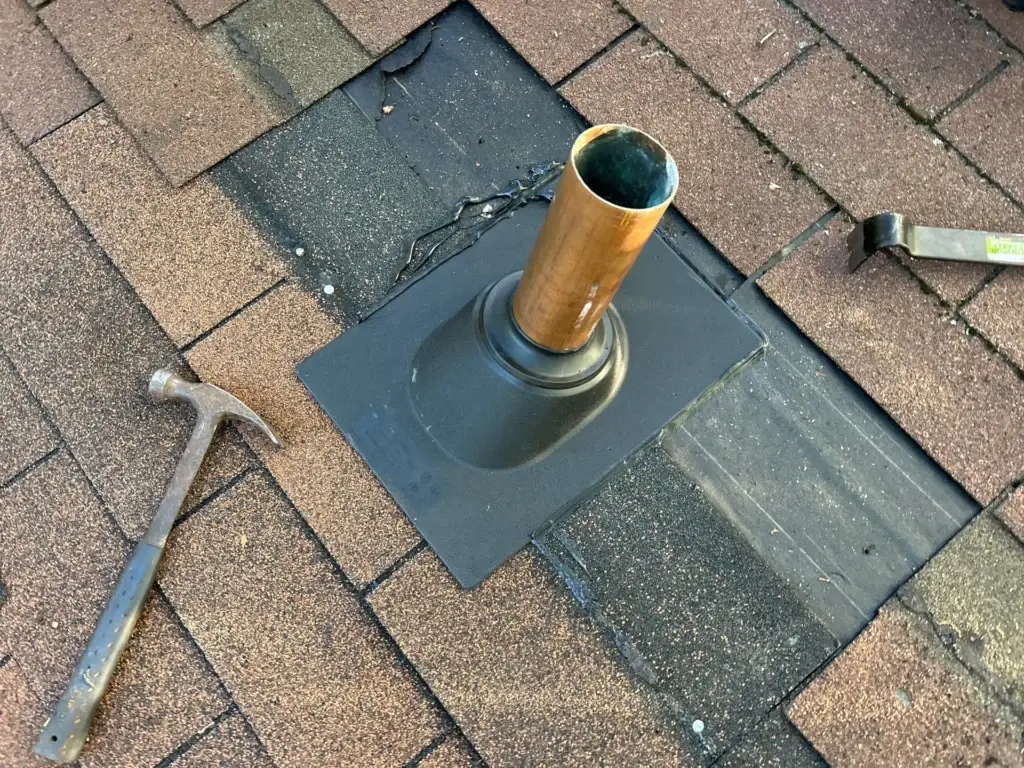 This image shows a pipe boot repair on a shingled roof. The old boot has been removed, and a new one is being installed. A copper pipe protrudes from the center of the boot, which is surrounded by shingles. A hammer and a tool lie nearby, indicating that the repair is in progress. The new boot appears to be securely fastened to the roof, ensuring a watertight seal around the pipe. Overall, the image suggests a professional and thorough repair job to prevent future leaks and damage to the roof.