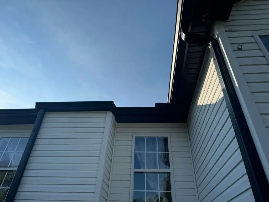 Newly installed black gutters adorn the exterior of a white house, complemented by a matching black downspout. The gutters are seamlessly integrated into the house's design, blending functionality with aesthetics. The image showcases the clean lines and modern look of the new gutter system, set against the crisp white siding of the house. The clear blue sky in the background adds a sense of freshness and completion to the scene, highlighting the successful installation of the new gutters.