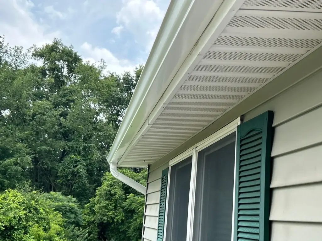 Close-up view of a newly installed white rain gutter system on a house, showcasing its seamless integration with the roof's edge and downspout. The gutter is attached to the side of the house, which features light green vinyl siding and a window with green shutters. The surrounding area is surrounded by lush green trees and a blue sky with white clouds, providing a serene backdrop for the gutter installation.