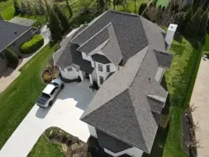 The image depicts a large, two-story house with a gray roof, showcasing a roof replacement project. The house features a white brick exterior, a chimney, and a covered front porch. A white truck is parked in the driveway, surrounded by a well-manicured lawn with green grass and various plants. The image is taken from an aerial perspective, likely from a drone, providing a clear view of the roof's condition and the surrounding property.