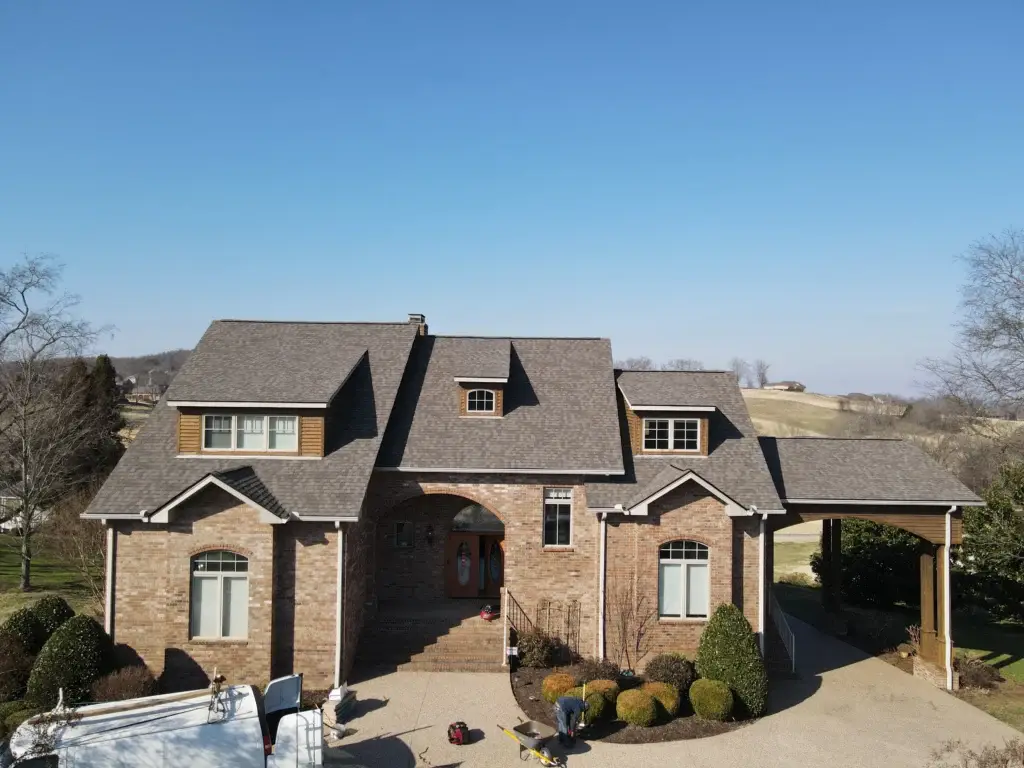 Recently completed shingle roof installation by Gold Medal Roofing: Aerial view of stunning two-story brick house with newly installed gray shingle roof, covered front porch, and expansive driveway. A white truck and roofing equipment indicate the finished project. Surrounded by lush trees and bushes, under a clear blue sky.