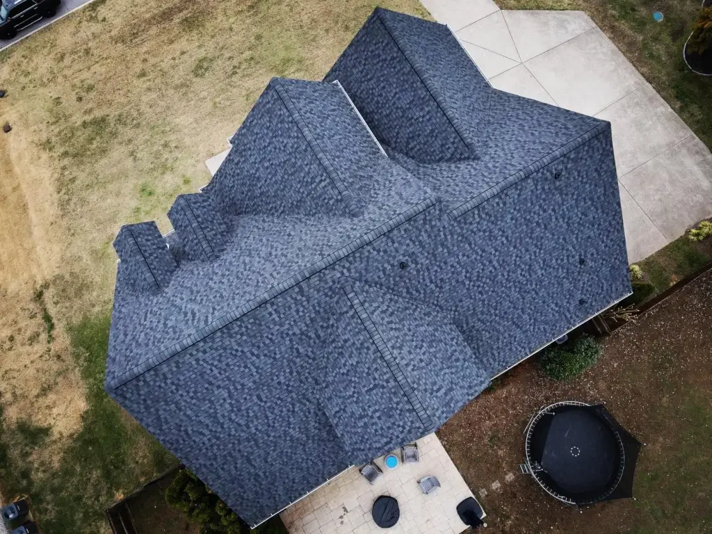 erial view of two-story house with sleek gray shingle roof, perfectly replaced for durability and curb appeal, complemented by crisp white walls. Lush backyard features patio seating, trampoline, vibrant garden, and surrounding fence. Idyllic setting for relaxation, showcasing expert shingle roof replacement.