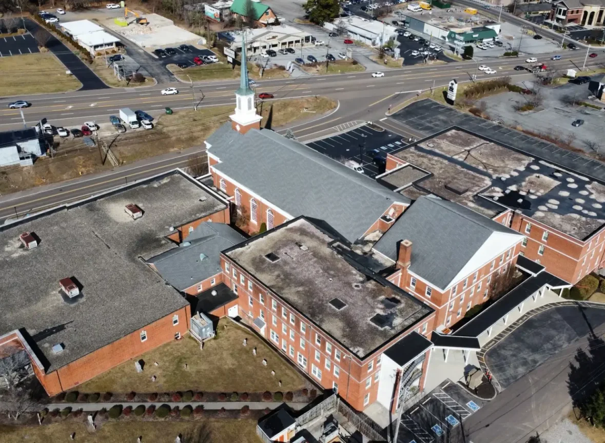 This aerial image showcases a newly installed roof on a large church building, featuring a striking grey color with white trim accents. The roof's design is characterized by multiple sections, including a central steeple and a smaller, rectangular section on the left. The surrounding landscape is dotted with other buildings, roads, and parking lots, providing context to the church's location. The image highlights the roof's clean lines, modern aesthetic, and attention to detail, making it an attractive addition to the church's architecture. The use of grey and white colors creates a sense of serenity and sophistication, while the multiple sections add visual interest and depth to the design. Overall, the image effectively showcases the newly installed roof as a beautiful and functional element of the church's structure.